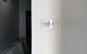 clear acrylic sign mounted with two silver standoffs that reads "115 ELECTRICAL CLOSET"