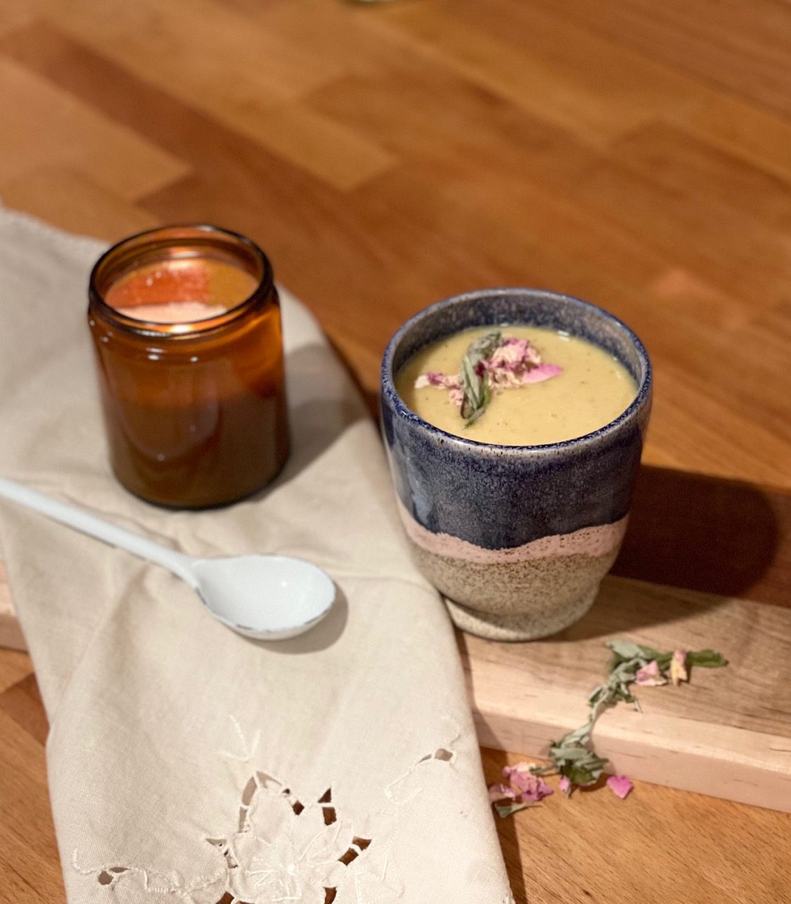 Handmade ceramic cup with mango mugwort mushroom milk adorned with dried rose petals and mugwort leaves, surrounded by cottage things: a warm wooden cutting board draped with an embroidered napkin, a lit candle in a warm amber jar, and scattered dried herbs