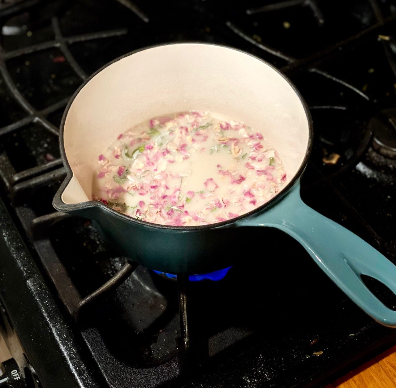 Teal cast-iron, enamel-covered saucepan with a handle simmering charmingly on a dark gas-powered stove top. Inside is oat milk and a colorful herbal blend of rose petals and mugwort