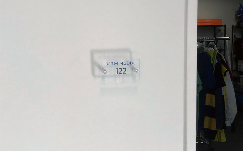 clear acrylic sign mounted with two silver standoffs that reads "XRM MEDIA 122"