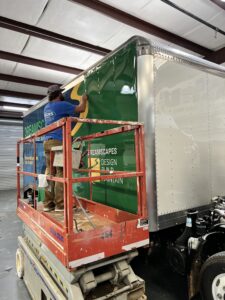 Installer applying green vinyl wrap to a box truck from a scissor lift at Signs & More’s shop in Marietta, GA